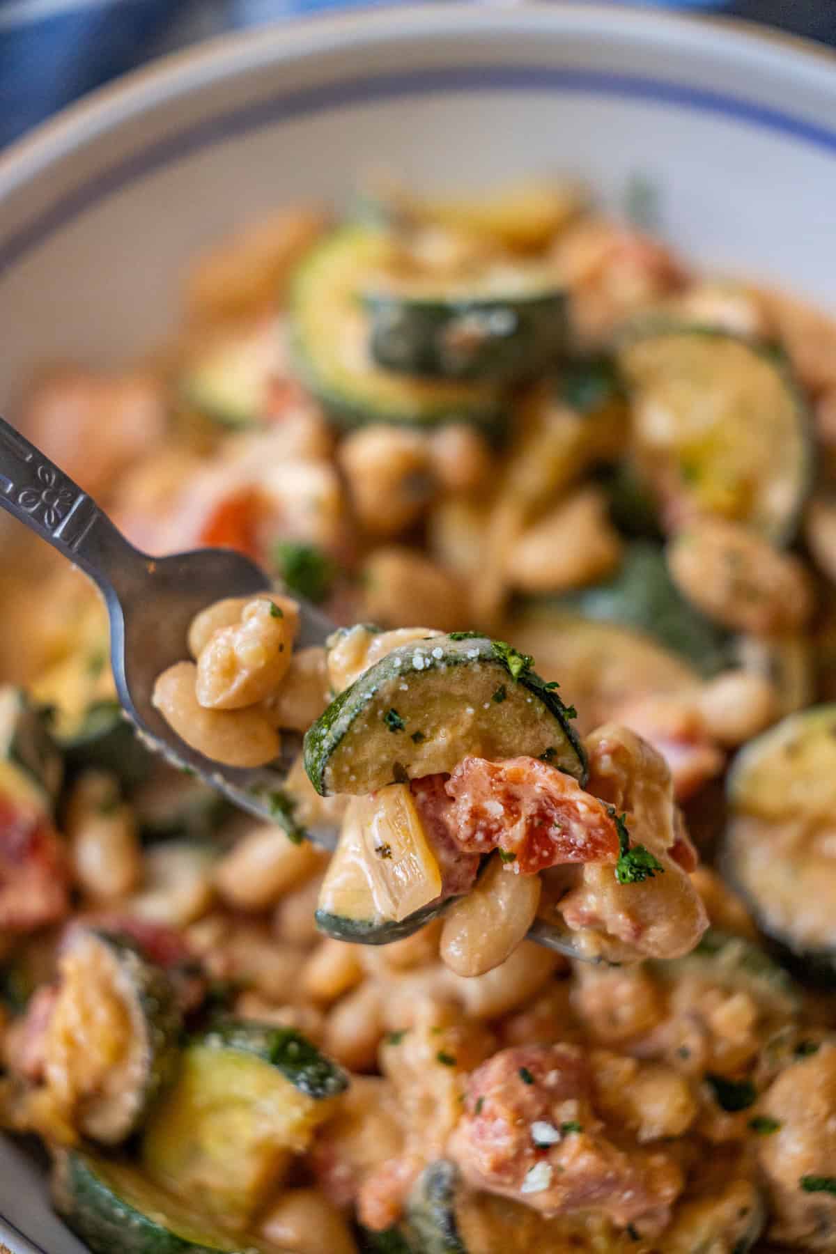 A close-up of a forkful of pasta dish featuring zucchini, diced tomatoes, white beans, and creamy sauce that looks so good it might make you say "marry me," with the rest of the dish blurred in the background.
