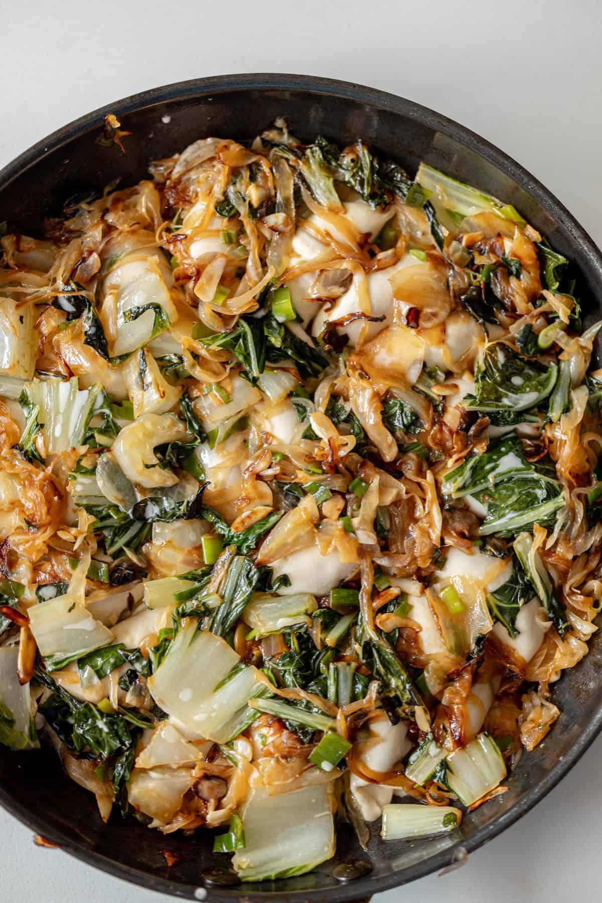 A close-up of a pan filled with cooked bok choy, onions, and melted cheese, reminiscent of a savory fried potsticker, on a light background.