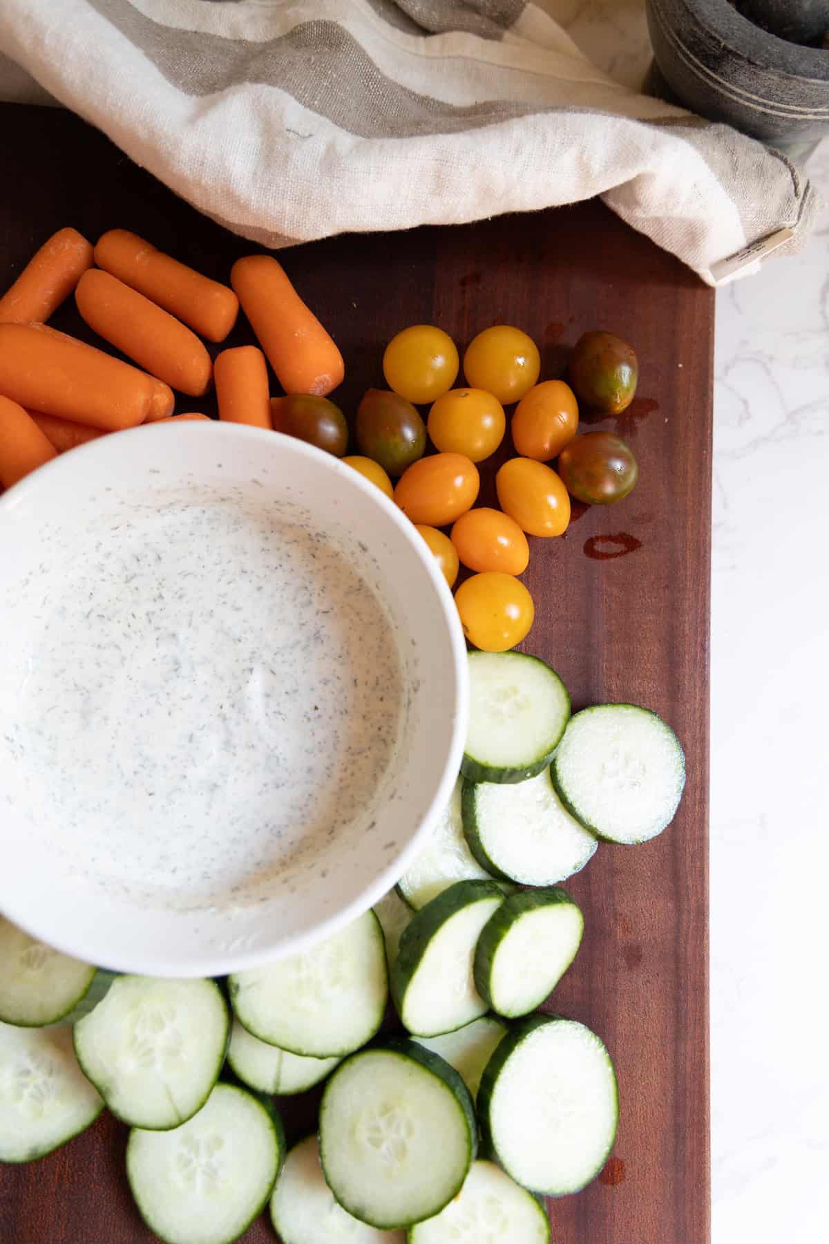 A bowl of creamy homemade ranch dressing sits on a wooden board, surrounded by sliced cucumbers, cherry tomatoes, and baby carrots, with a striped cloth in the background.