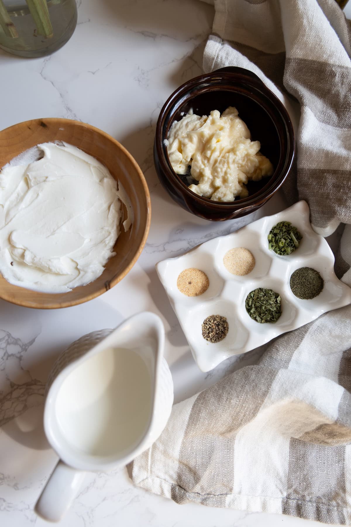 A wooden bowl of cream cheese, a brown bowl of mayonnaise, assorted dried herbs and spices, and a white jug of milk on a marble surface with a striped cloth—perfect for making homemade ranch dressing.