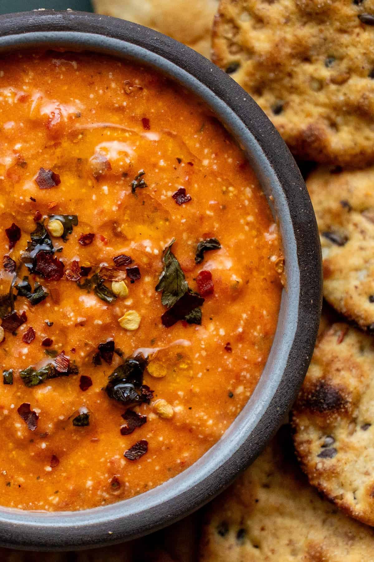 A bowl of creamy red dip garnished with herbs and chili flakes, surrounded by round, crispy crackers.