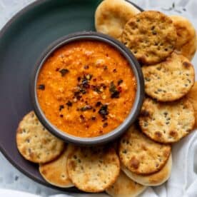 A bowl of red pepper dip garnished with chili flakes, surrounded by round crackers and mini flatbreads on a dark plate.
