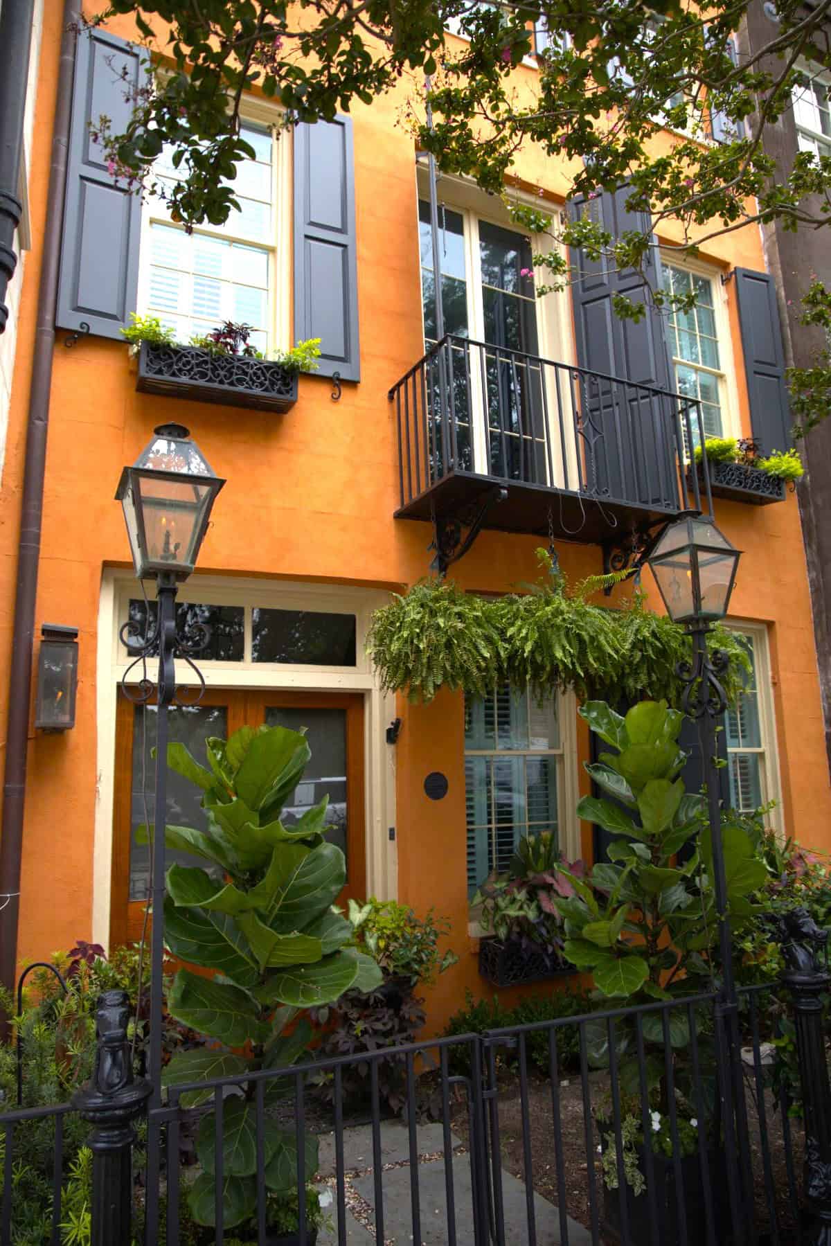Three-story orange townhouse with black shutters and balcony, window flower boxes, lush greenery, and a black iron fence in front—perfect for any Charleston travel guide.