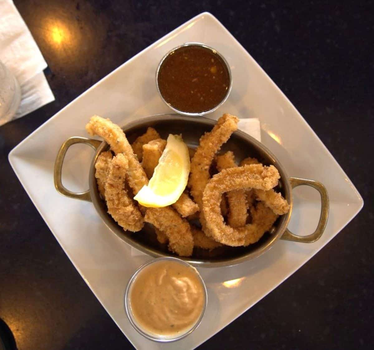 A white plate with breaded fried strips, a lemon wedge, and two dipping sauces in metal cups, photographed from above—perfect for any Charleston travel guide foodie adventure.