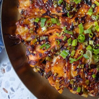 Close-up of a savory dish featuring sliced potatoes topped with chopped green onions, sesame seeds, and a spicy, caramelized sauce on a gold plate—served alongside crispy potsticker pancakes.