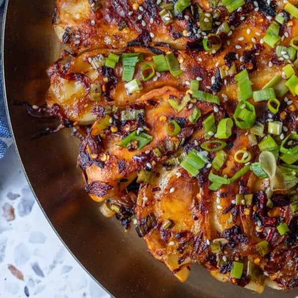 Close-up of a savory dish featuring sliced potatoes topped with chopped green onions, sesame seeds, and a spicy, caramelized sauce on a gold plate—served alongside crispy potsticker pancakes.