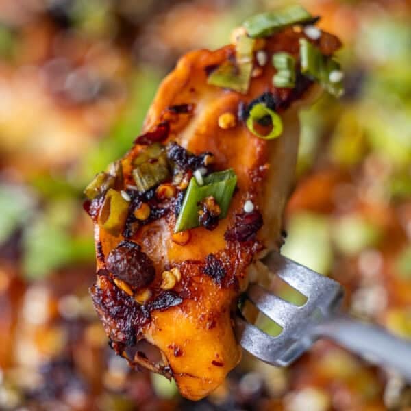 A close-up of a fork holding a piece of cooked chicken topped with chopped green onions, red pepper flakes, and sesame seeds, inspired by our Spicy Potsticker Crunch Cake Recipe.