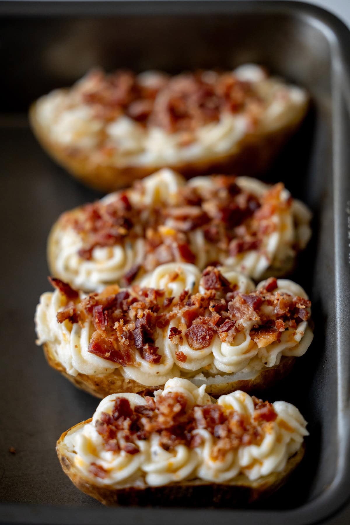 Four loaded potato halves, twice baked and topped with piped mashed potatoes and crumbled bacon, are arranged in a baking pan.