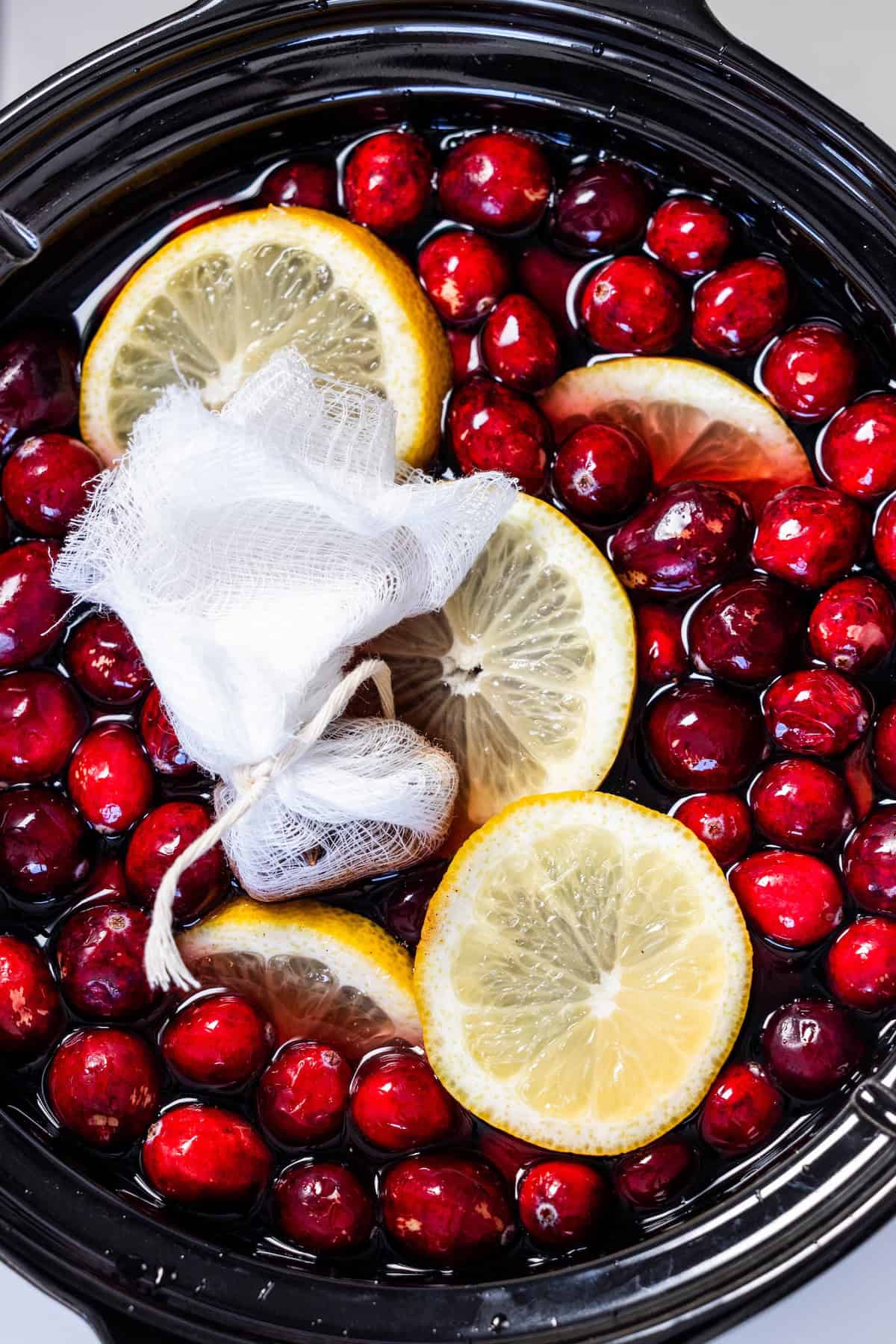 A black pot filled with fresh cranberries, lemon slices, and a tied cheesecloth bundle soaking in liquid—perfect for making homemade Mulled Cranberry Cider.