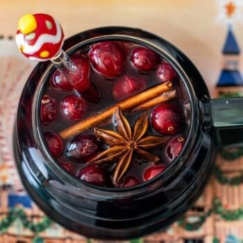 A glass teapot filled with Mulled Cranberry Cider, whole cranberries, two cinnamon sticks, a star anise, and a decorative stir stick.