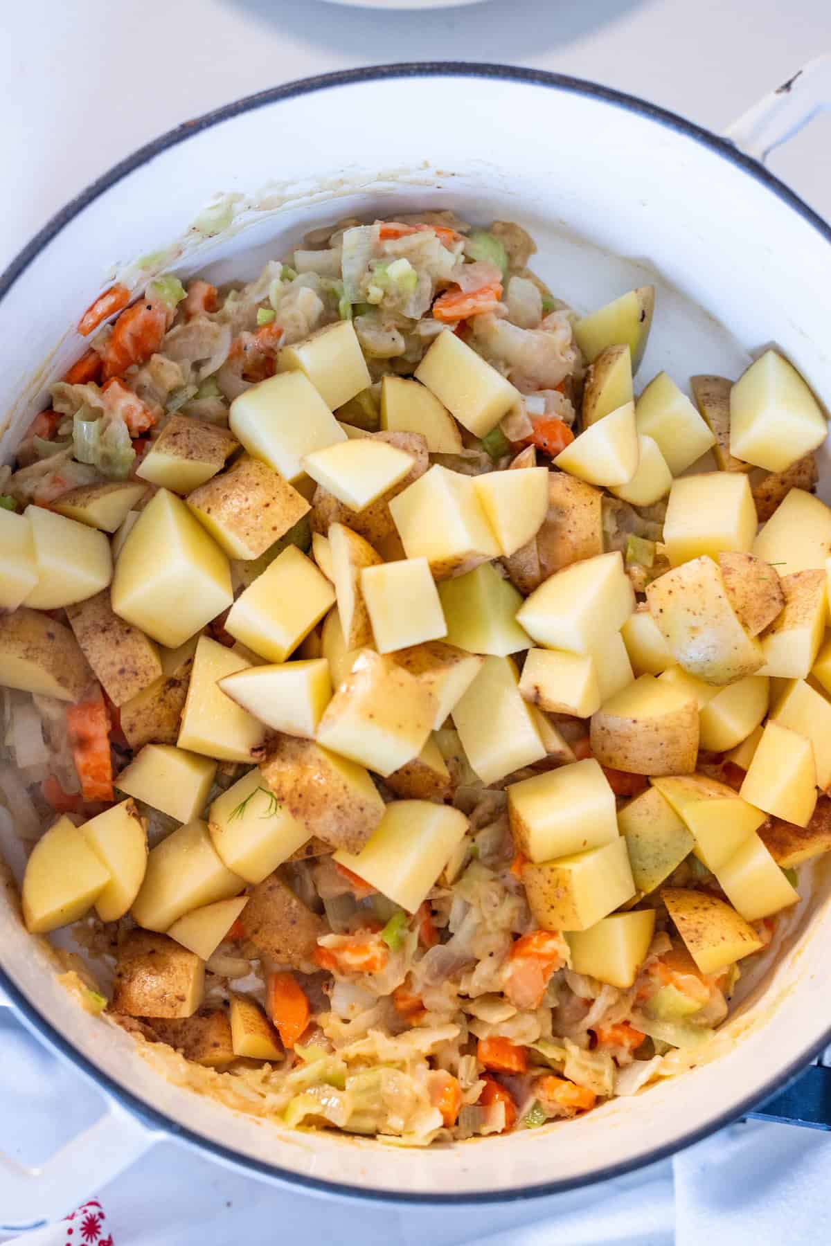 A white pot filled with chopped potatoes, carrots, and cabbage, some of the vegetables appear to be sautéed, evokes the comforting base of a traditional Irish Chowder.