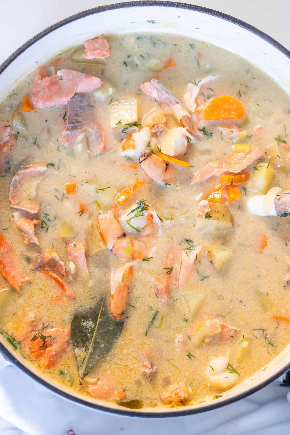 A pot of creamy Irish Seafood Chowder with salmon pieces, shrimp, diced potatoes, carrots, herbs, and a bay leaf, being stirred with a wooden spoon.