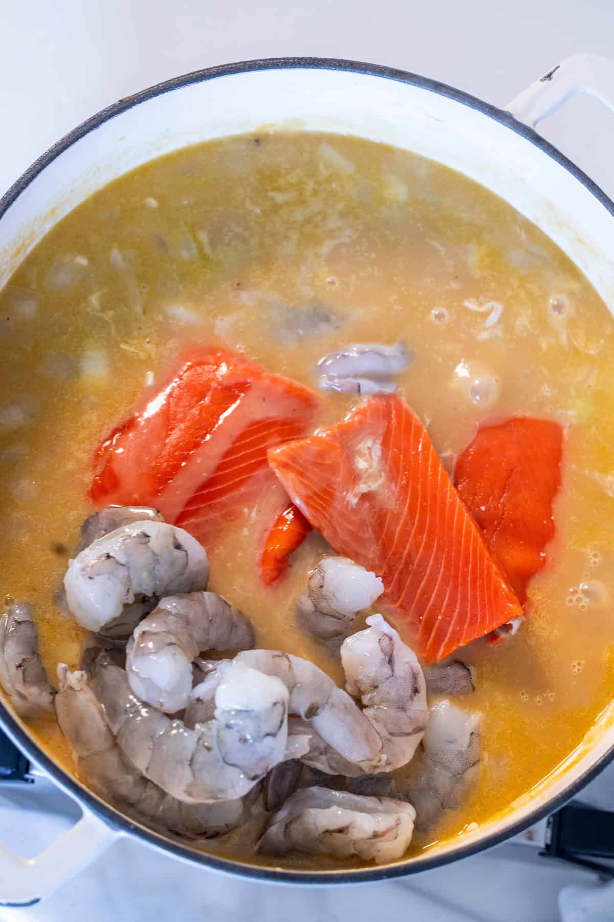A pot of Irish Seafood Chowder with raw salmon fillets and shrimp in a light broth, shown simmering on the stovetop.
