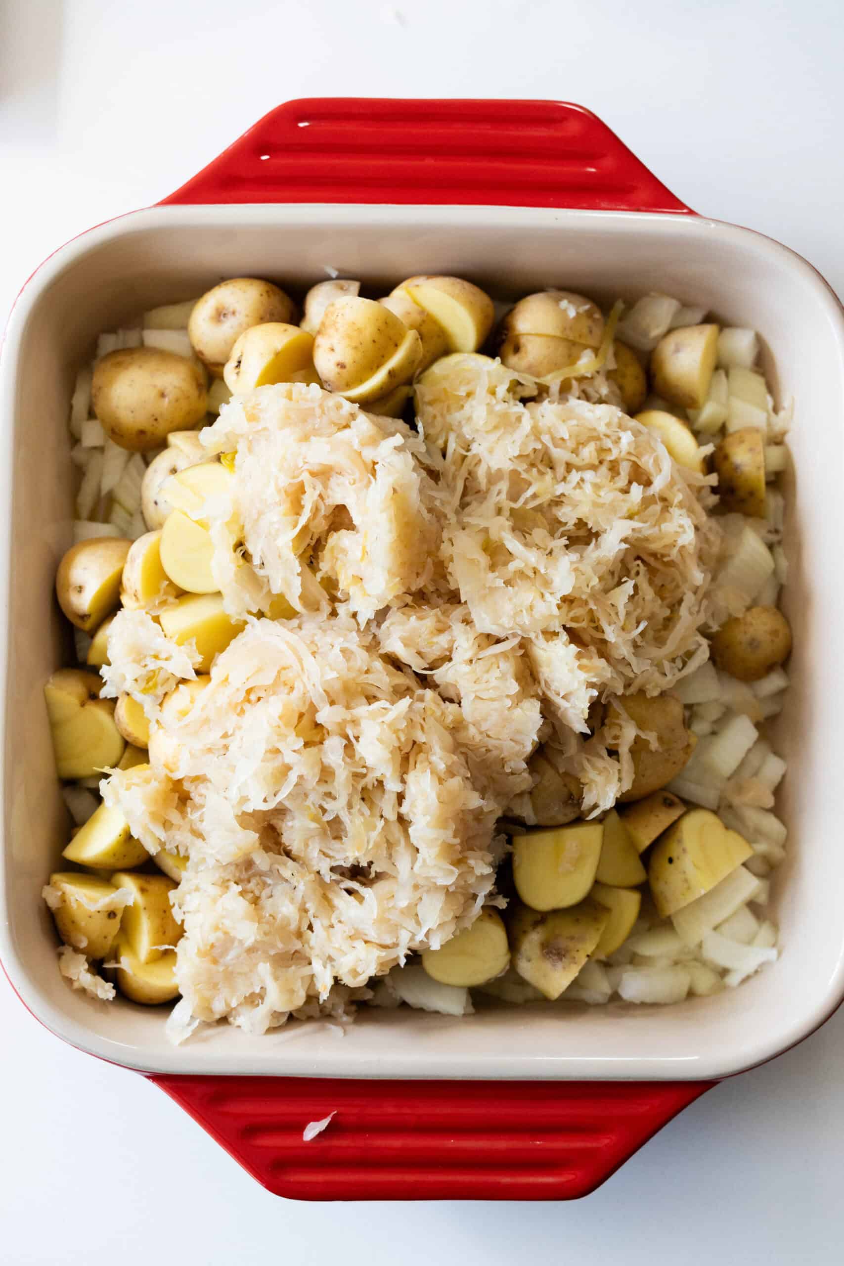 A red baking dish filled with chopped roasted potatoes, diced onions, and a layer of sauerkraut on top, ready for cooking.