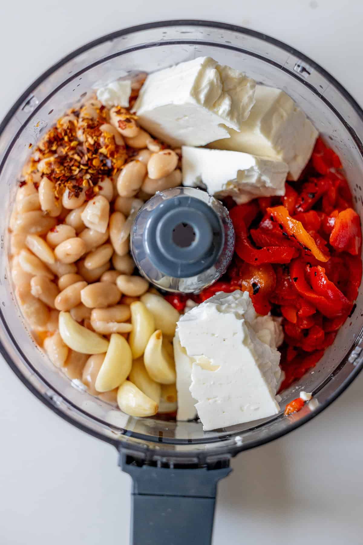 Food processor bowl containing white beans, garlic cloves, feta cheese, roasted red peppers, and red pepper flakes, viewed from above before mixing.