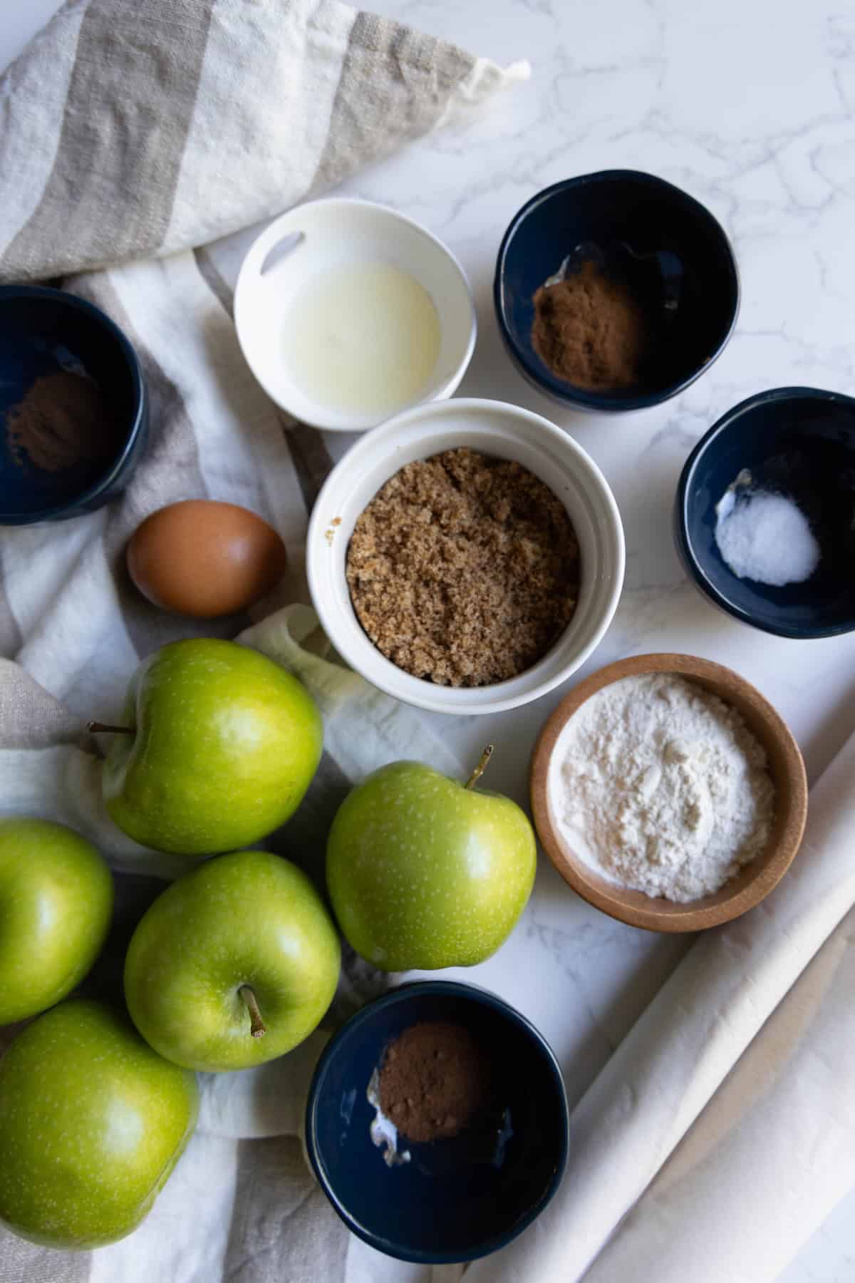 Overhead view of baking ingredients for apple pie, including green apples, brown sugar, flour, an egg, spices, baking powder, and a small bowl of liquid on a white surface with a striped towel.