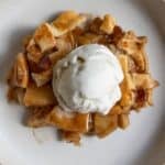 A slice of apple pie with pieces of crust is topped with a scoop of vanilla ice cream on a white plate.