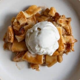 A slice of apple pie with pieces of crust is topped with a scoop of vanilla ice cream on a white plate.