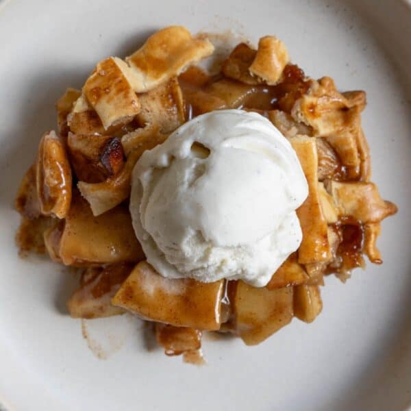 A slice of apple pie with pieces of crust is topped with a scoop of vanilla ice cream on a white plate.