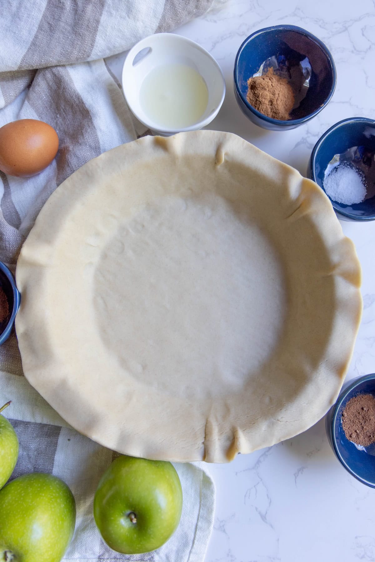Unbaked pie crust in a pie dish surrounded by bowls of ingredients, a whole egg, and green apples on a white surface—perfect for creating a classic apple pie.