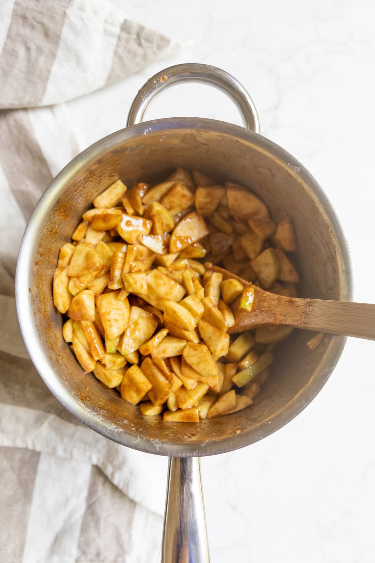 Sliced apples mixed with apple pie spices in a saucepan, stirred with a wooden spoon, on a light surface with a striped cloth nearby.