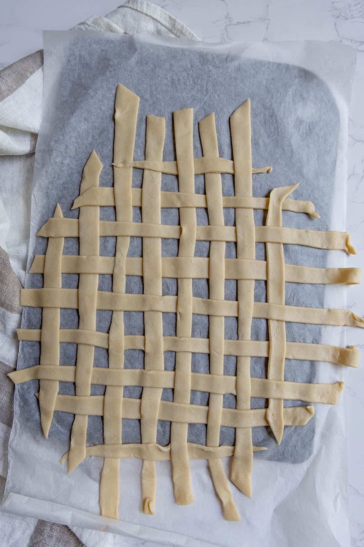 Unbaked lattice pie crust strips, perfect for an apple pie, are arranged in a grid pattern on parchment paper atop a baking sheet.