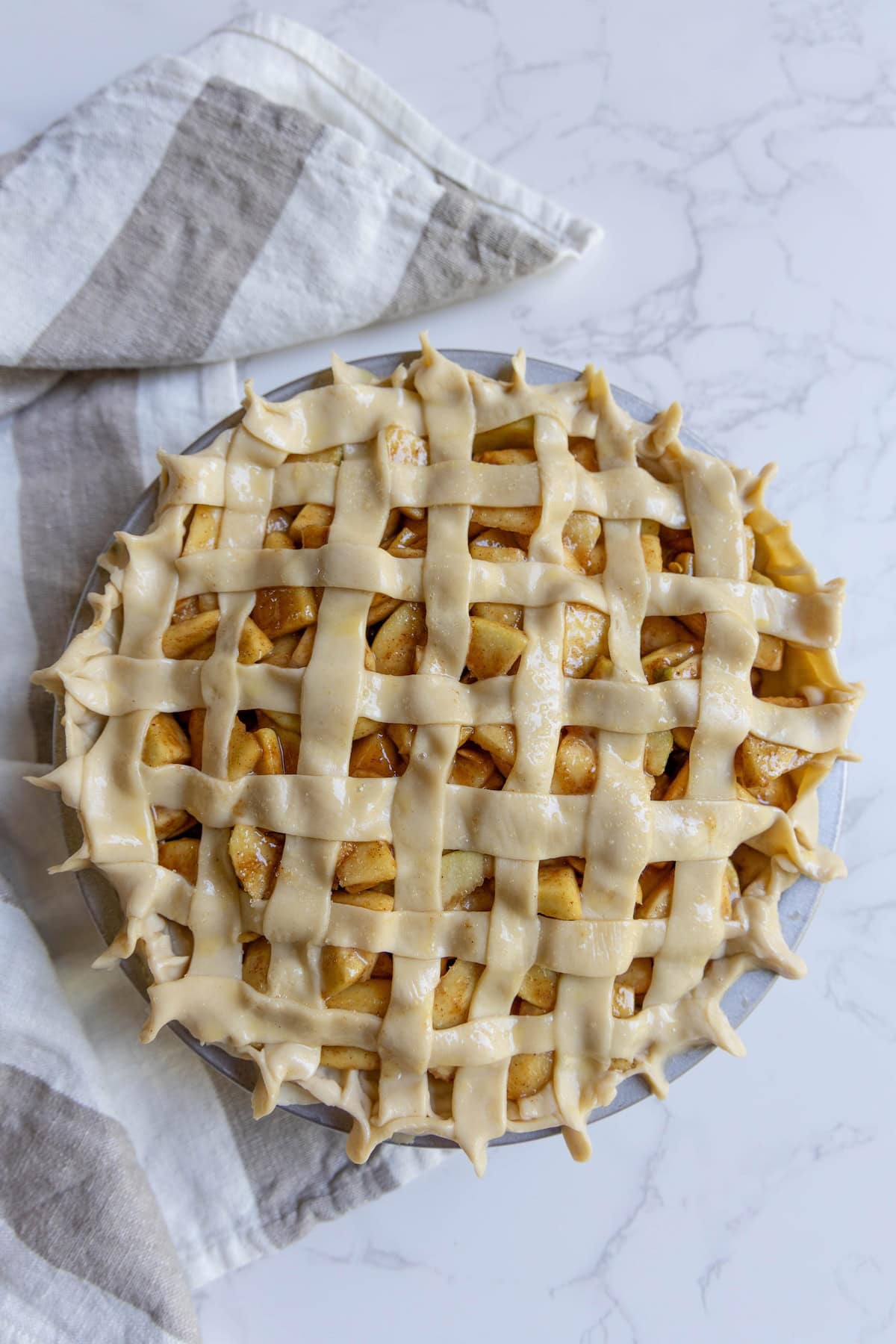 Unbaked apple pie with a lattice crust sits on a marble surface, next to a striped kitchen towel, ready to become a classic homemade apple pie.