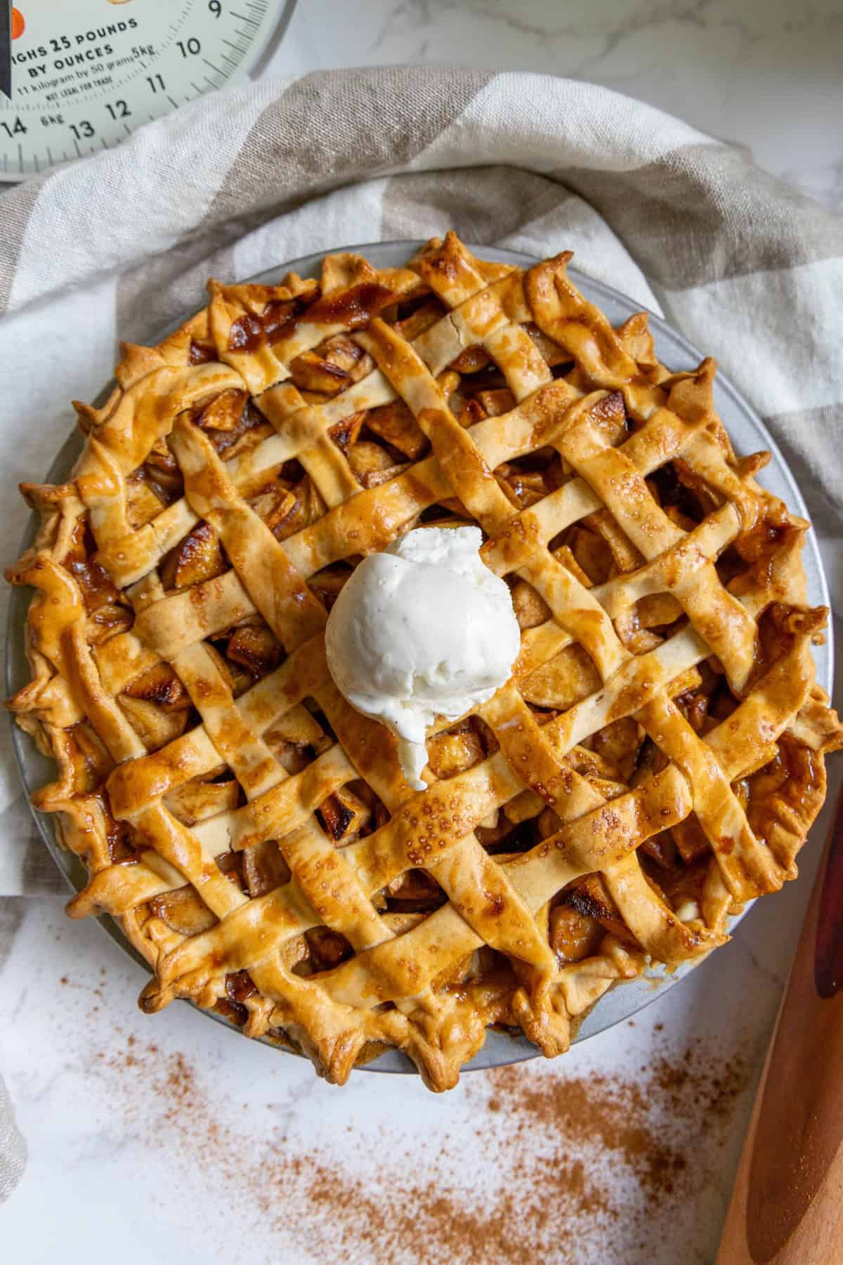 A classic baked apple pie with a lattice crust, topped with vanilla ice cream, rests on a striped kitchen towel, accompanied by a scale and a dusting of cinnamon.
