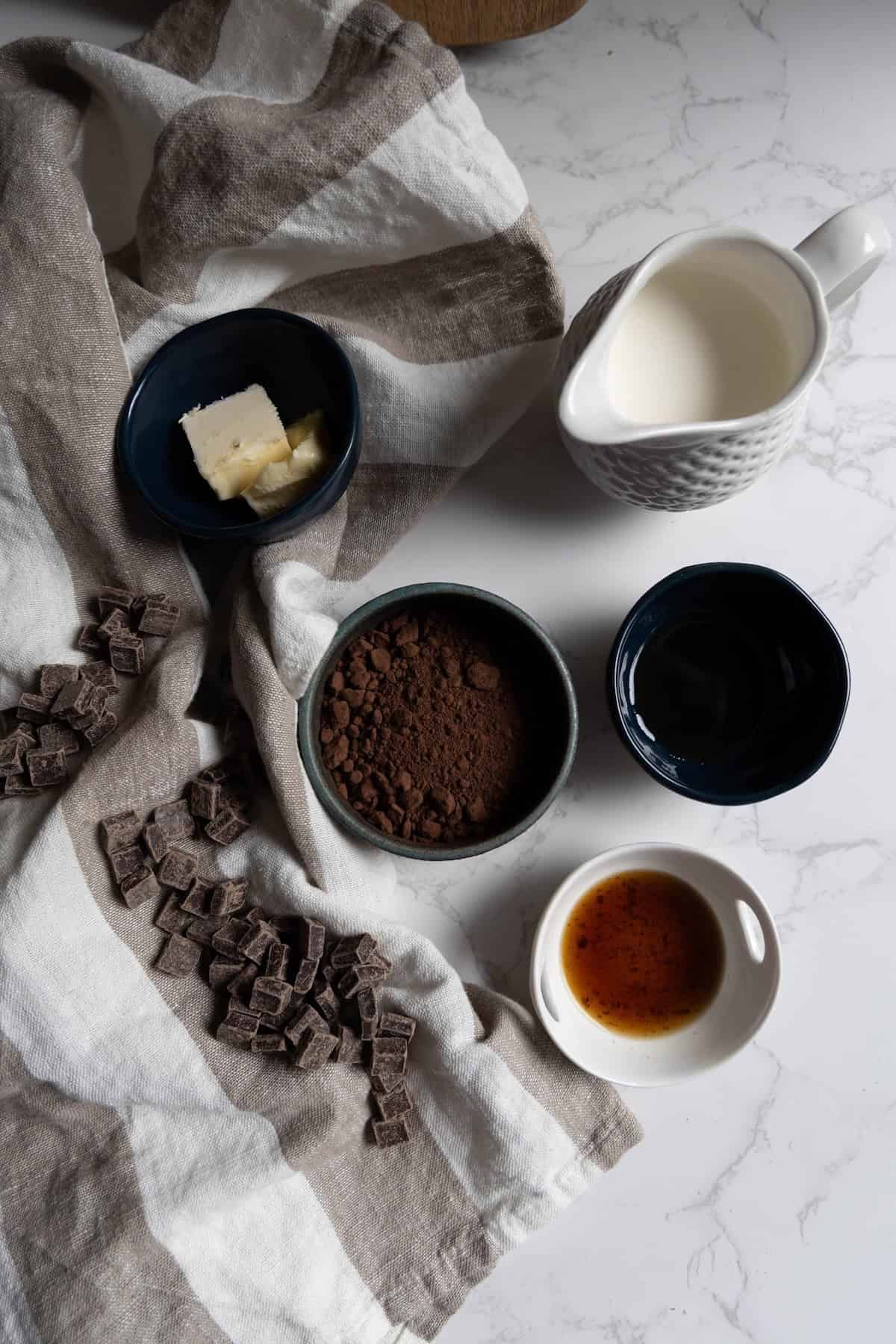 Several small bowls with butter, cocoa powder, cream, chocolate chunks, water, and vanilla extract—perfect ingredients for chocolate truffles—are arranged on a striped cloth on a white marble surface.
