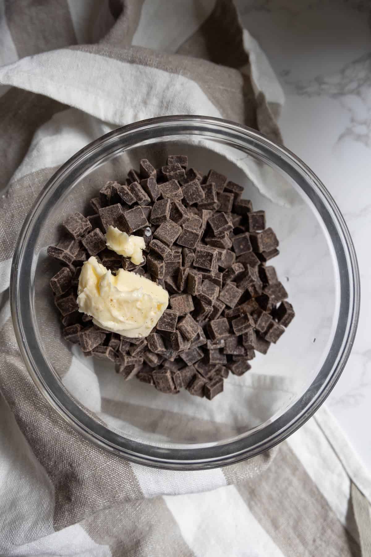 A glass bowl filled with chocolate chunks and a dollop of butter, perfect for making rich chocolate truffles, sits atop a striped cloth.