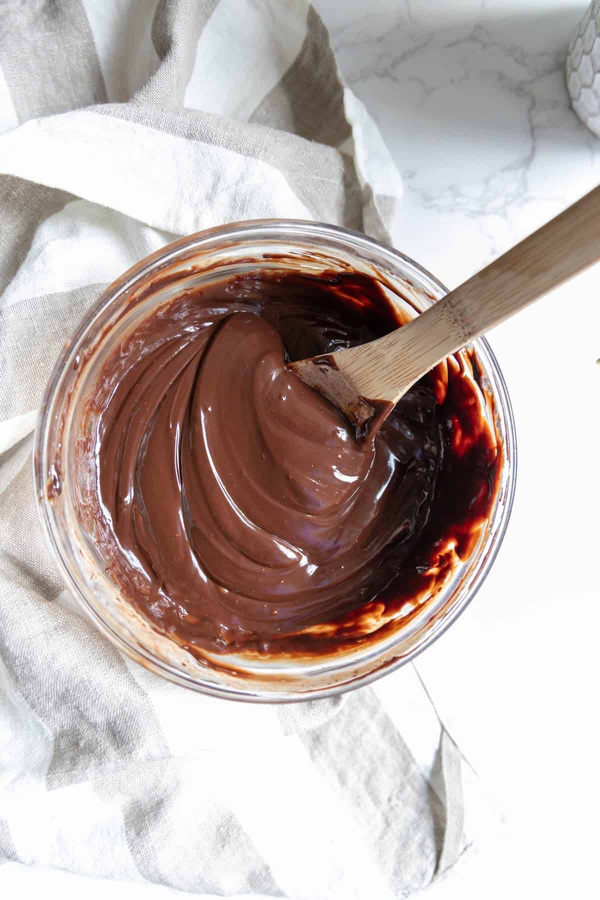 A glass bowl of smooth, glossy chocolate mixture for chocolate truffles sits with a wooden spoon resting inside, placed on a striped cloth.