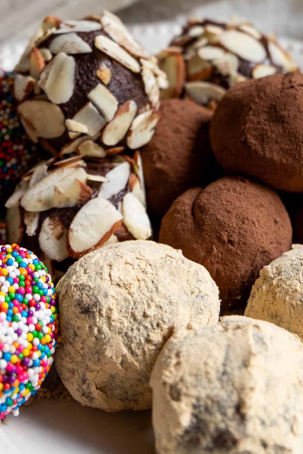 Assorted chocolate truffles are covered in sliced almonds, cocoa powder, powdered sugar, and colorful sprinkles, all beautifully arranged on a plate.