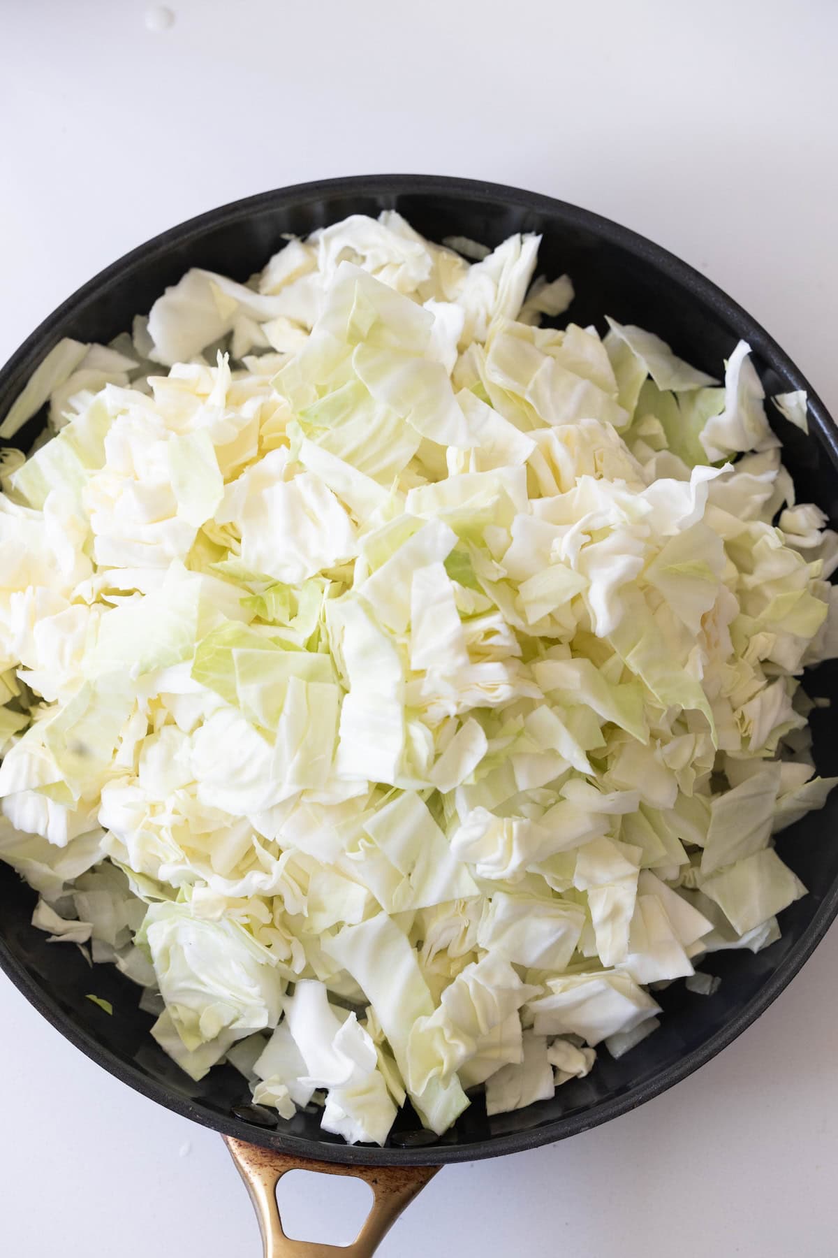 Chopped raw cabbage piled in a black skillet on a white surface, ready to be cooked for a classic cabbage and noodles dish.