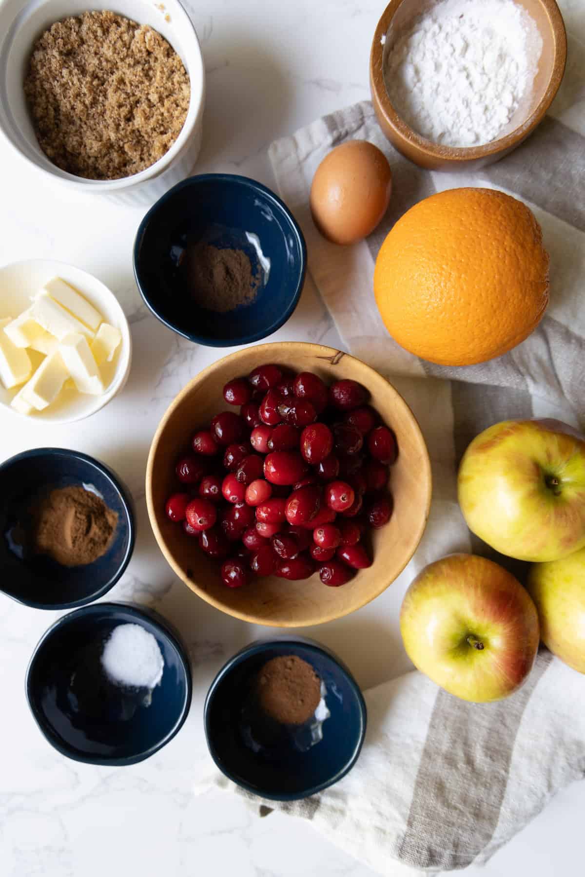 A top-down view of baking ingredients for cranberry apple pie, including cranberries, apples, an orange, butter, an egg, brown sugar, powdered sugar, and spices in bowls on a white surface.