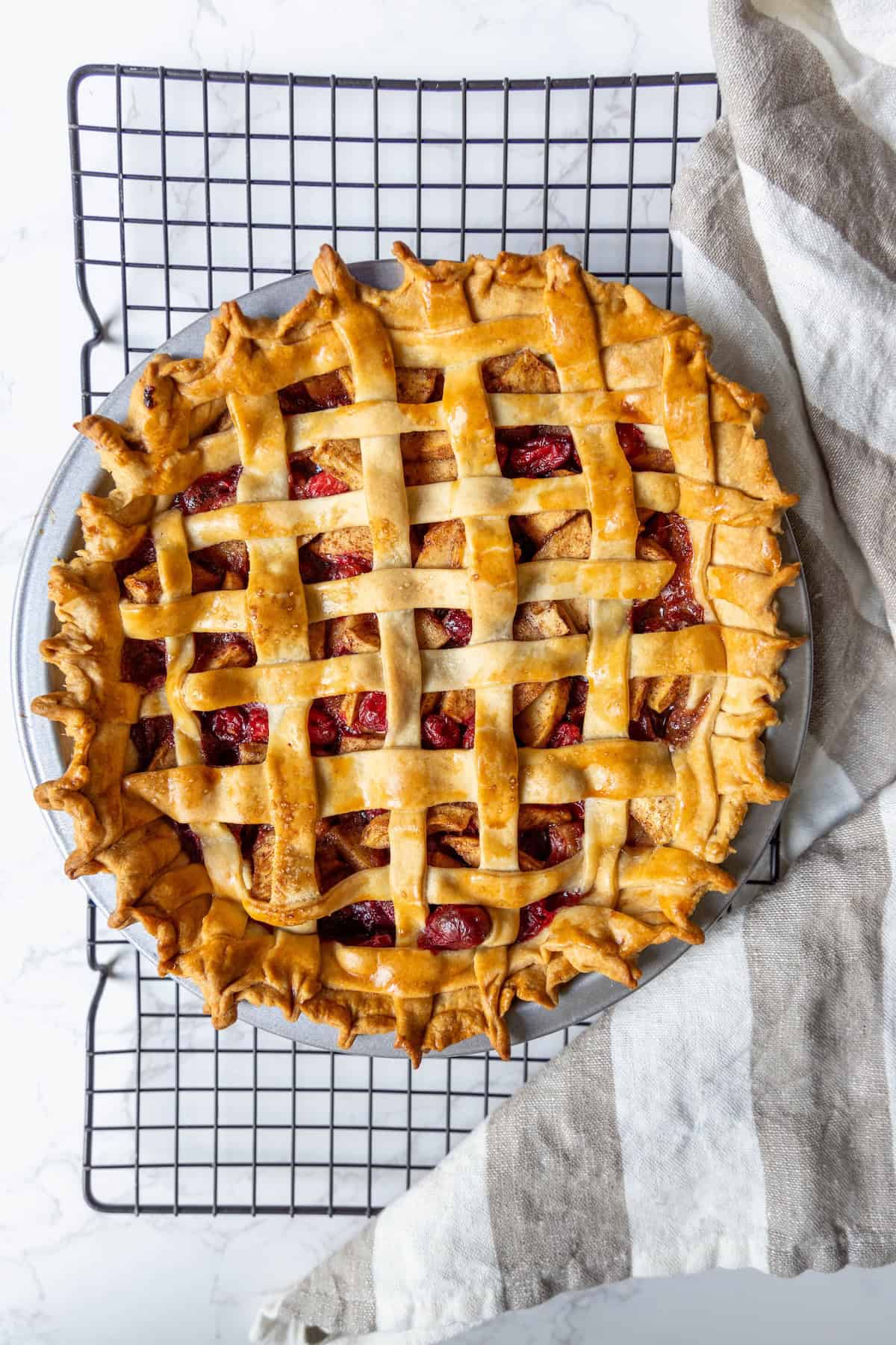 A freshly baked cranberry apple pie with a golden lattice crust sits on a cooling rack next to a striped cloth.
