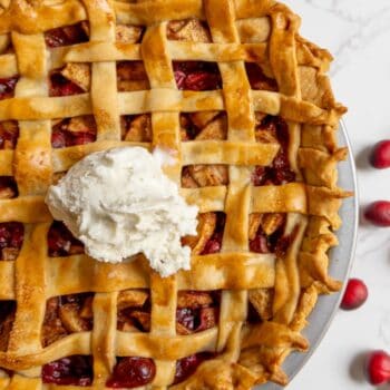 A lattice-topped cranberry apple pie with a scoop of vanilla ice cream in the center, placed on a white surface with a few cranberries nearby.