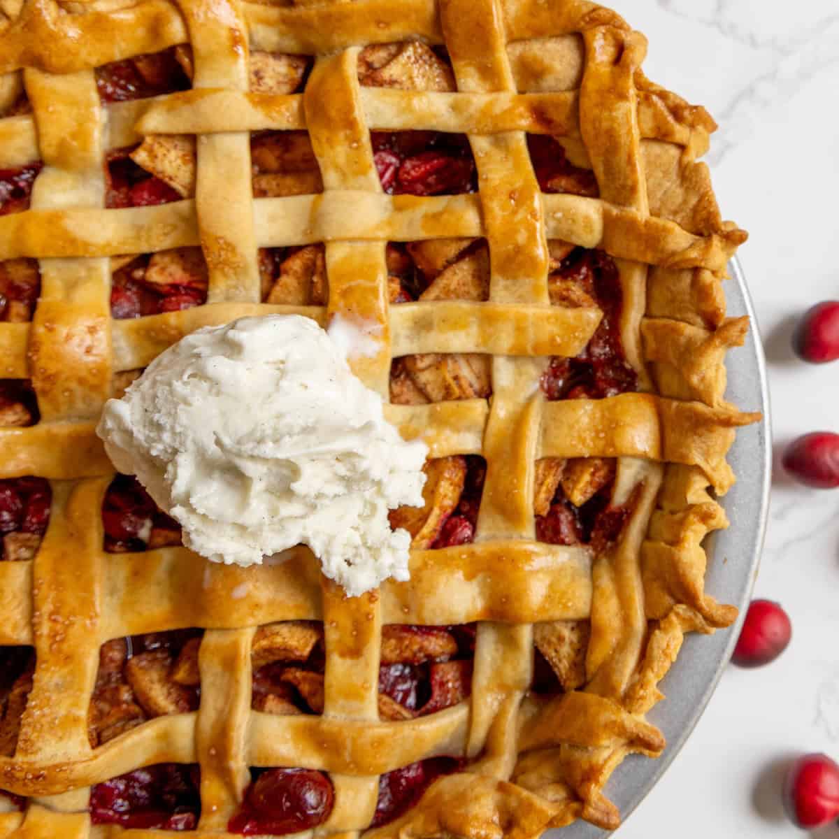 A lattice-topped cranberry apple pie with a scoop of vanilla ice cream in the center, placed on a white surface with a few cranberries nearby.