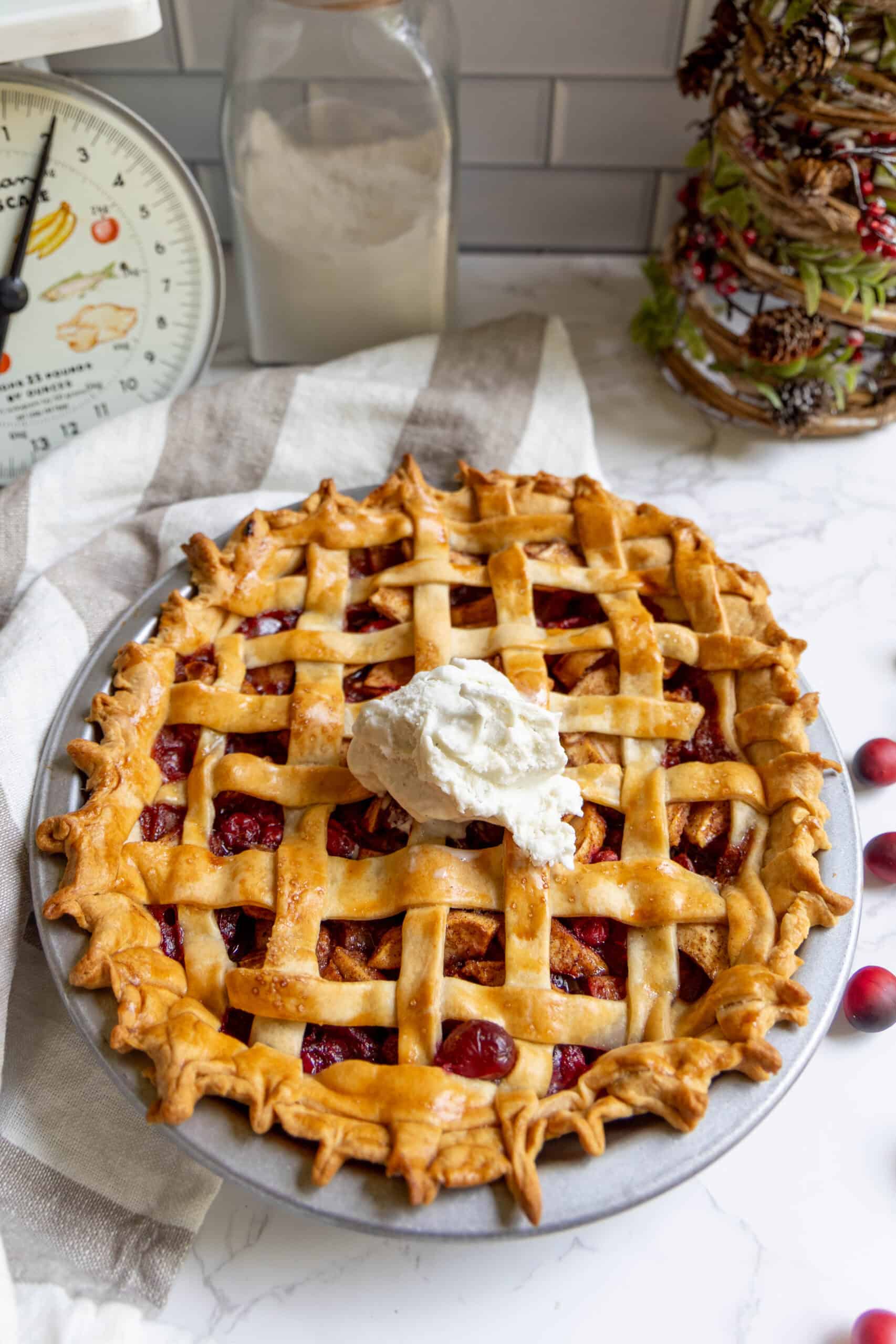 A lattice-topped cranberry apple pie with a dollop of whipped cream sits on a plate; kitchen items and cranberries are nearby on a marble countertop.