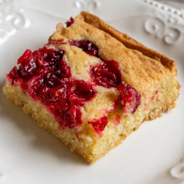 A square piece of yellow cake, reminiscent of cranberry bars, topped with baked cranberries on a white plate with decorative edges.