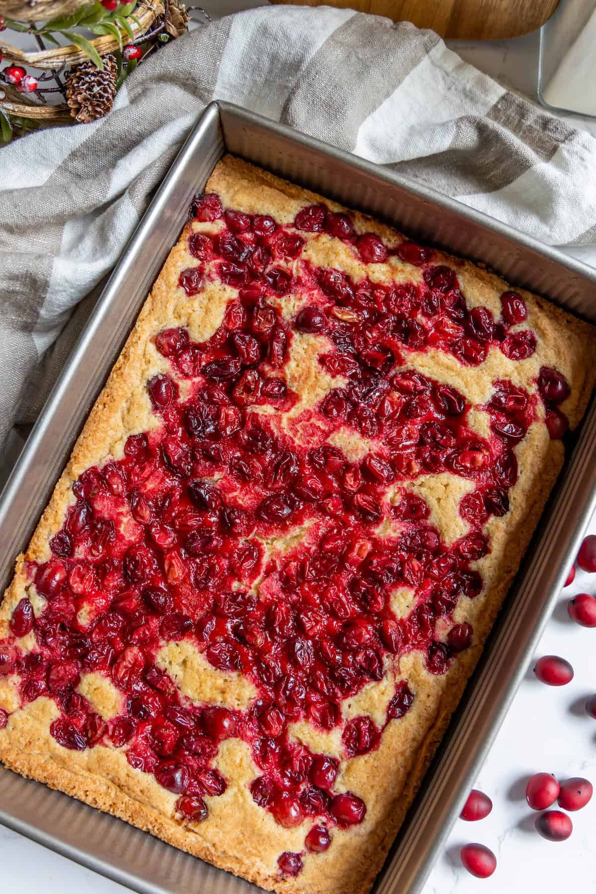 A rectangular baking pan with freshly baked cranberry bars sits on a cloth, topped with cranberries and surrounded by scattered fresh cranberries.