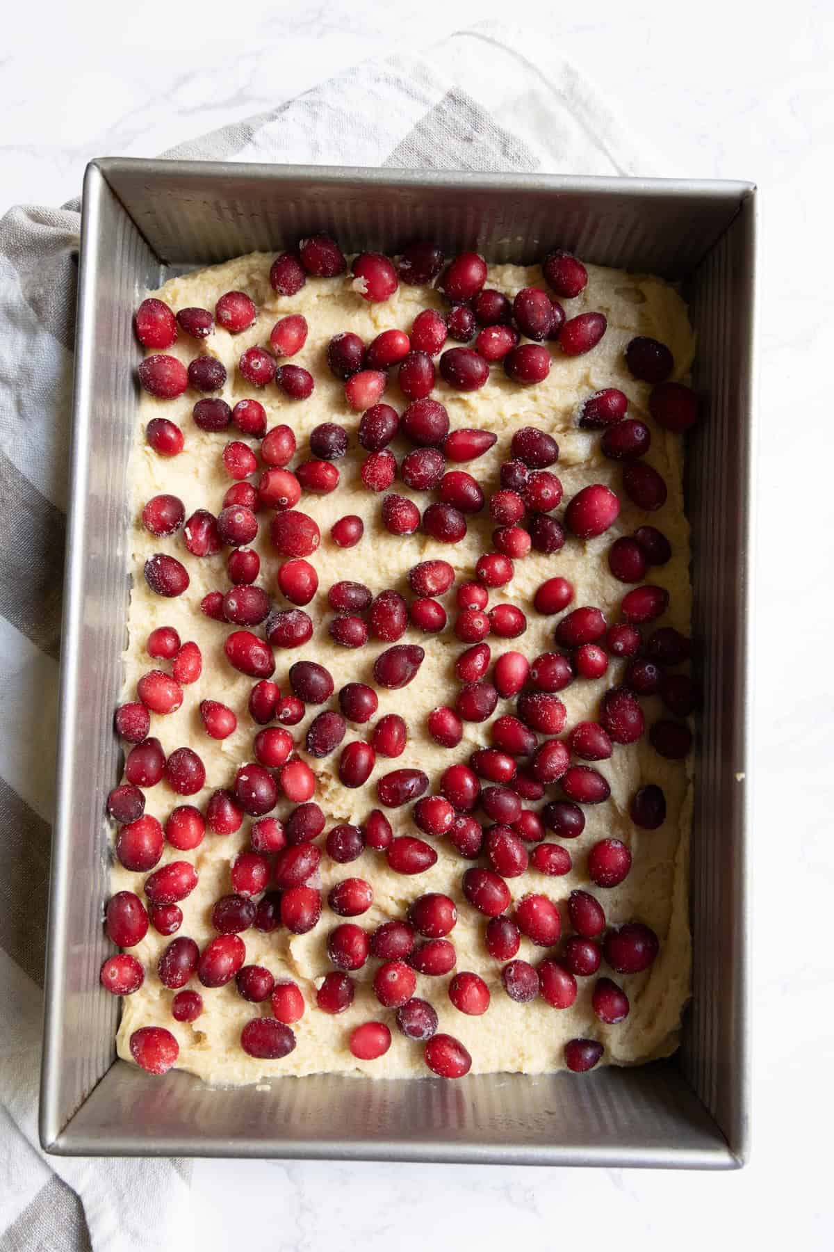A rectangular metal baking pan filled with unbaked cranberry bars dough topped with fresh red cranberries, placed on a striped kitchen towel.