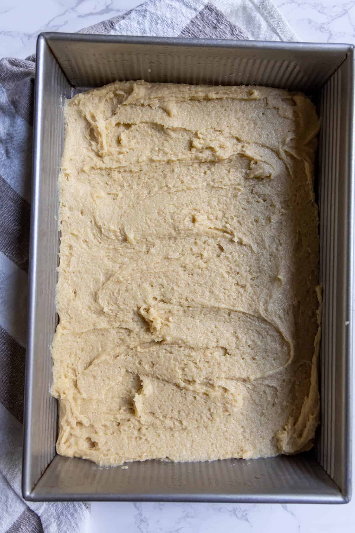 Rectangular metal baking pan filled with an even layer of raw cranberry bars dough, resting on a folded kitchen towel.