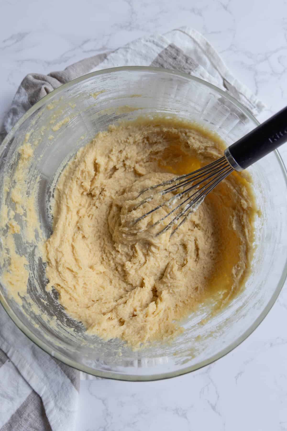 A glass bowl with pale, creamy batter for cranberry bars being mixed with a whisk, placed on a light-colored cloth and marble surface.