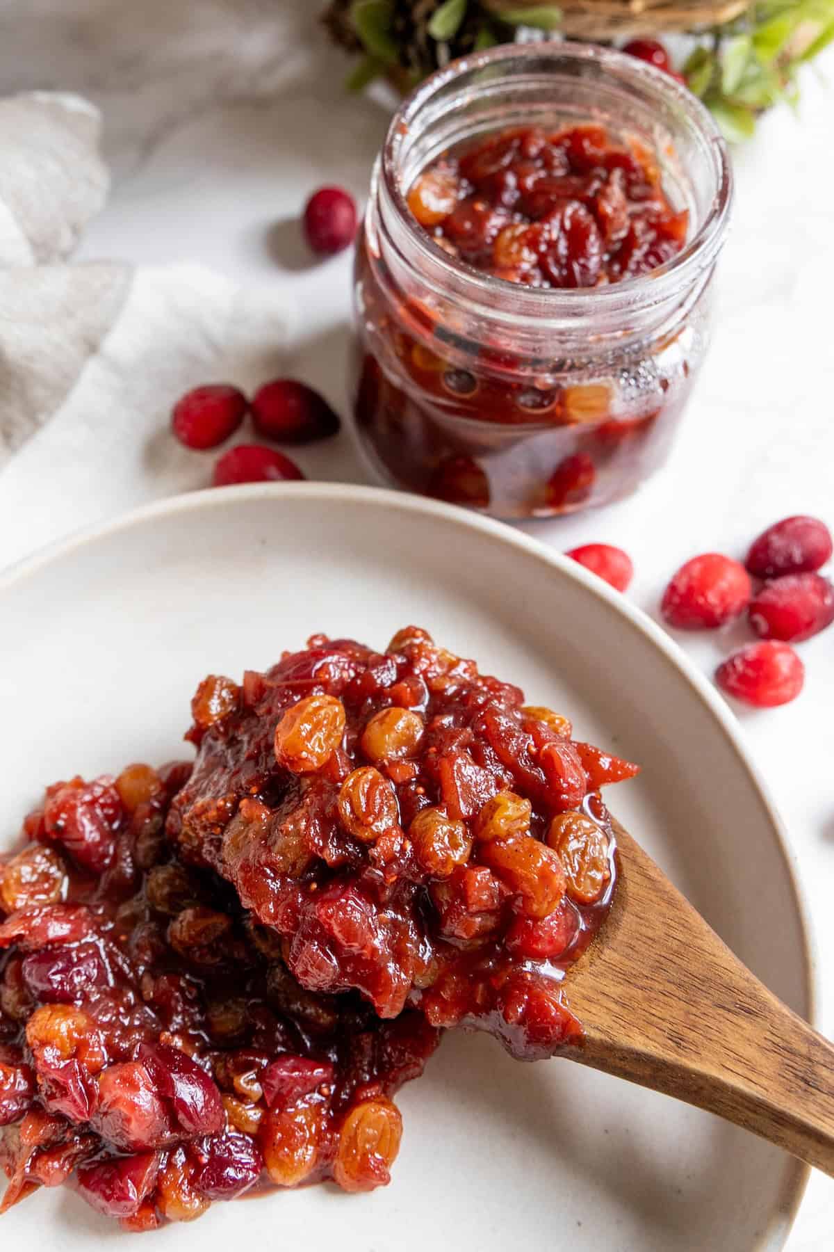 A wooden spoon holds a serving of cranberry chutney with raisins on a white plate, while more cranberry chutney sits in an open jar surrounded by scattered cranberries in the background.