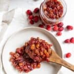 A wooden spoon holds a serving of cranberry chutney with raisins on a white plate, next to a jar of cranberry chutney and scattered fresh cranberries.