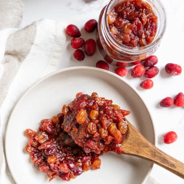 A wooden spoon holds a serving of cranberry chutney with raisins on a white plate, next to a jar of cranberry chutney and scattered fresh cranberries.