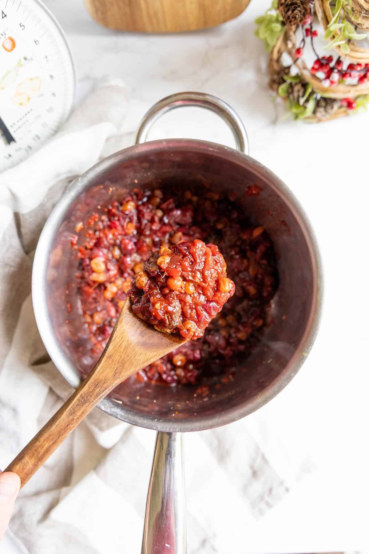 A wooden spoon holds up a portion of chunky, tomato-based cranberry chutney with beans over a stainless steel pot on a marble countertop.