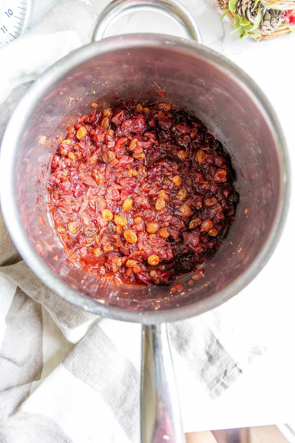 A stainless steel pot containing a cranberry chutney made with cooked dried fruits like raisins and cranberries, set on a white surface with a striped cloth partially visible.