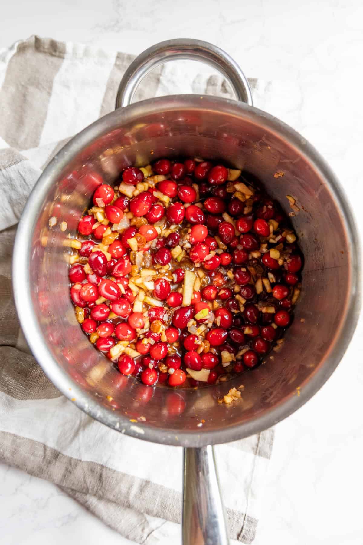 A stainless steel pot filled with cranberries, chopped onions, and seasonings sits on a striped kitchen towel, ready to be transformed into delicious cranberry chutney.