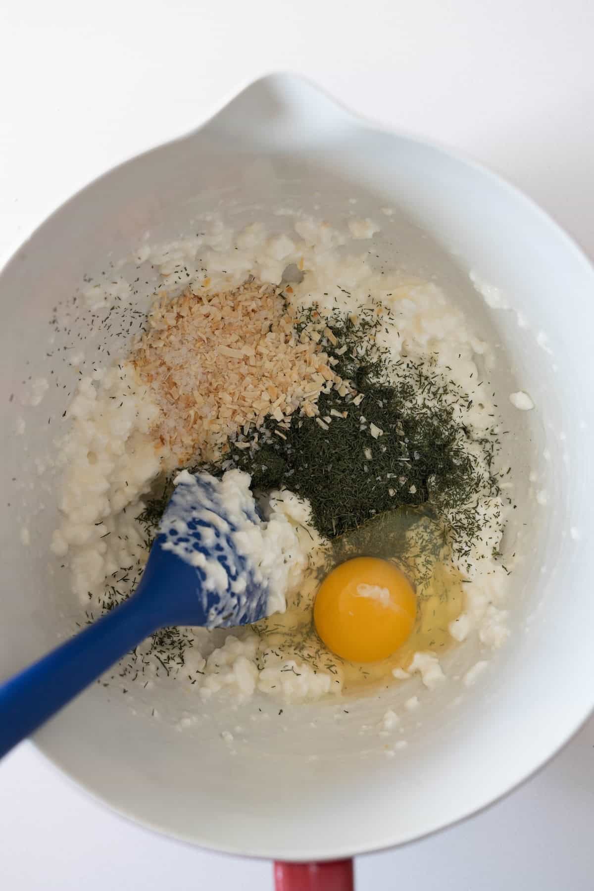 A mixing bowl containing cottage cheese, a raw egg, dried dill, dried onion flakes, and a blue spatula—perfect ingredients for whipping up a classic Dilly Bread Recipe.